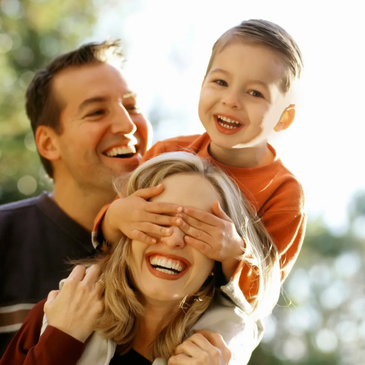 Parents laughing outdoors with their young son on their shoulders